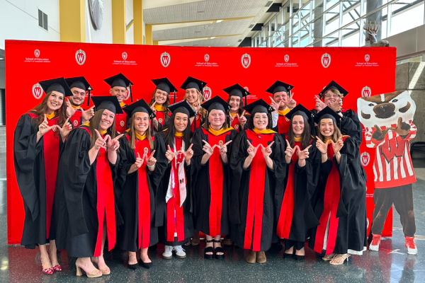 Graduates from the MS in Educational Psychology: Professional Educators (MSPE) Class of 2026 wearing graduation gowns and making the “W” hand signs in front of a canvas featuring the UW–Madison logo.