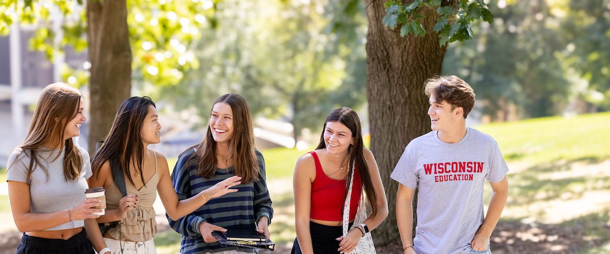 A group of 5 students walk on a sunny path with green leafy trees in the background