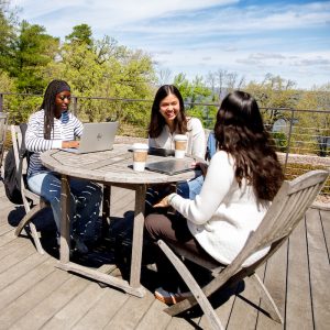 3 students study outdoors at a table on a rooftop while trees and blues skies are in the background